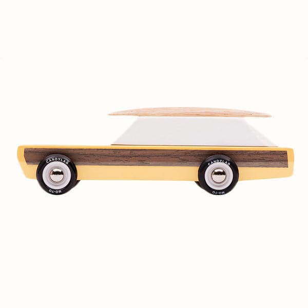 This wooden station wagon model has an elongated daffodil yellow body with walnut veneer-paneled sides, providing the ultimate classic feel. Wheels are jet black reading 'Candylab Mo-To' around white and silver rims. The windowed top half consists of a single white trapezoid shaped wooden block. A magnetic beech wood surfboard lays directly on top.